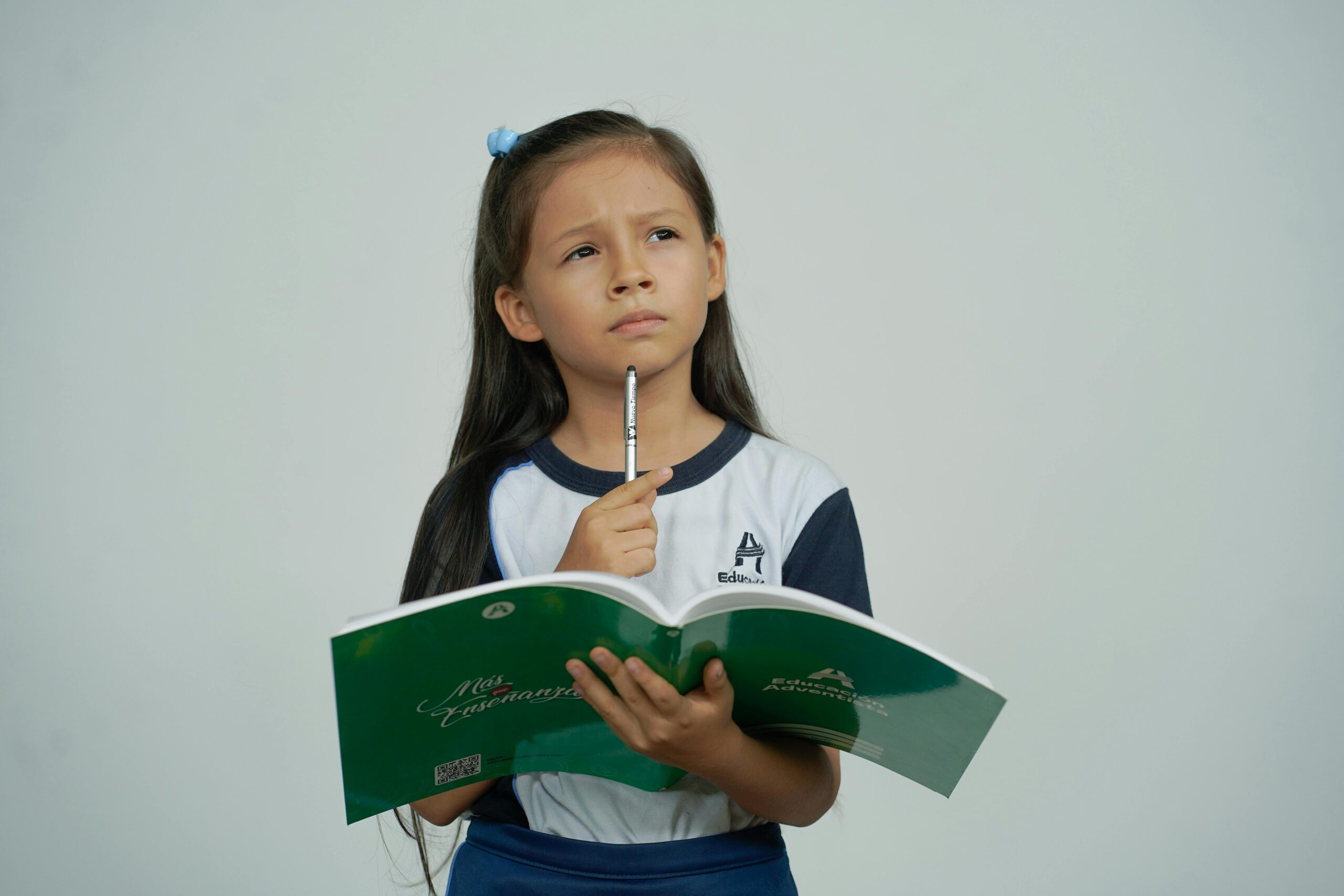 A young girl holds a green notebook, appearing thoughtful in a study setting.